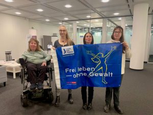 Von links nach rechte: Dunja Robin, Karin Winklhofer, Umaahan Gräsle, Konstanze Kobell stehen in einem Büro und halten ein Banner vor sich hoch. Darauf steht: "Frei leben - ohne Gewalt". Das Banner ist dunkelblau und von Terre des Femmes. Es ist das Banner zum Aktionsmonat gegen Gewalt an Frauen. Es ist in Gelb der Umriss einer Frau gemalt, welche in einer Befreiungsgeste die Arme weit offen nach oben hält; ihr Haar weht hinter ihr.