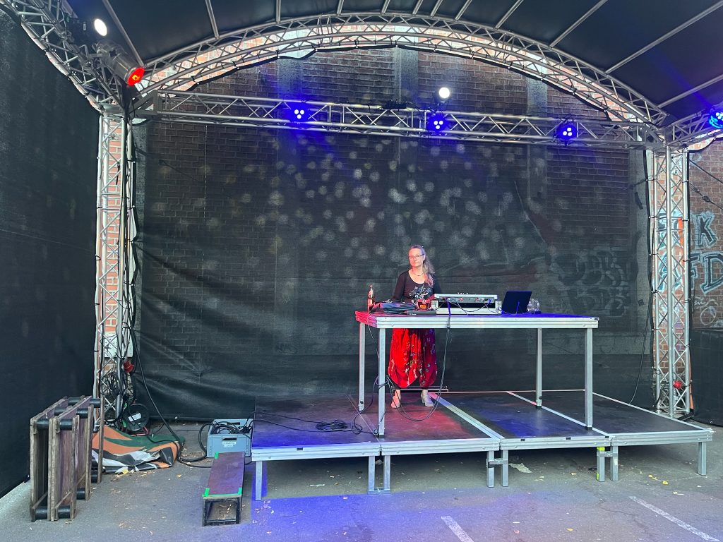 Karin Winklhofer steht am DJ-Pult auf einer überdachten Bühne vor der mit Graffiti besprühten Ziegelwand des Kulturhauses und wird von blauen Scheinwerfern angestrahlt.