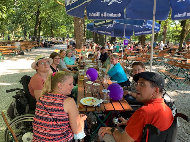 Frauen (teilweise mit Rollstuhl) sitzen mit einigen Männern im Biergarten an einem langen Tisch zusammen. Schirme sorgen für Schatten, lila Luftballons mit Netzwerkfrauen-Bayern-Aufdruck sind auf den Tischen.