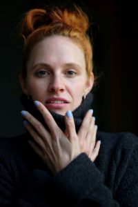 Bild von Kasandra Wedel mit einem schwarzen Rollkragenpullover bekleidet; die Hände sind überkreuzt bis fast zum Gesicht erhoben, Handflächen zum Betrachter. Die Fingernägel sind hellblau lackiert.