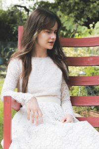 Sängerin CassMae in einem weißen Kleid im Garten auf einem Holzstuhl sitzend