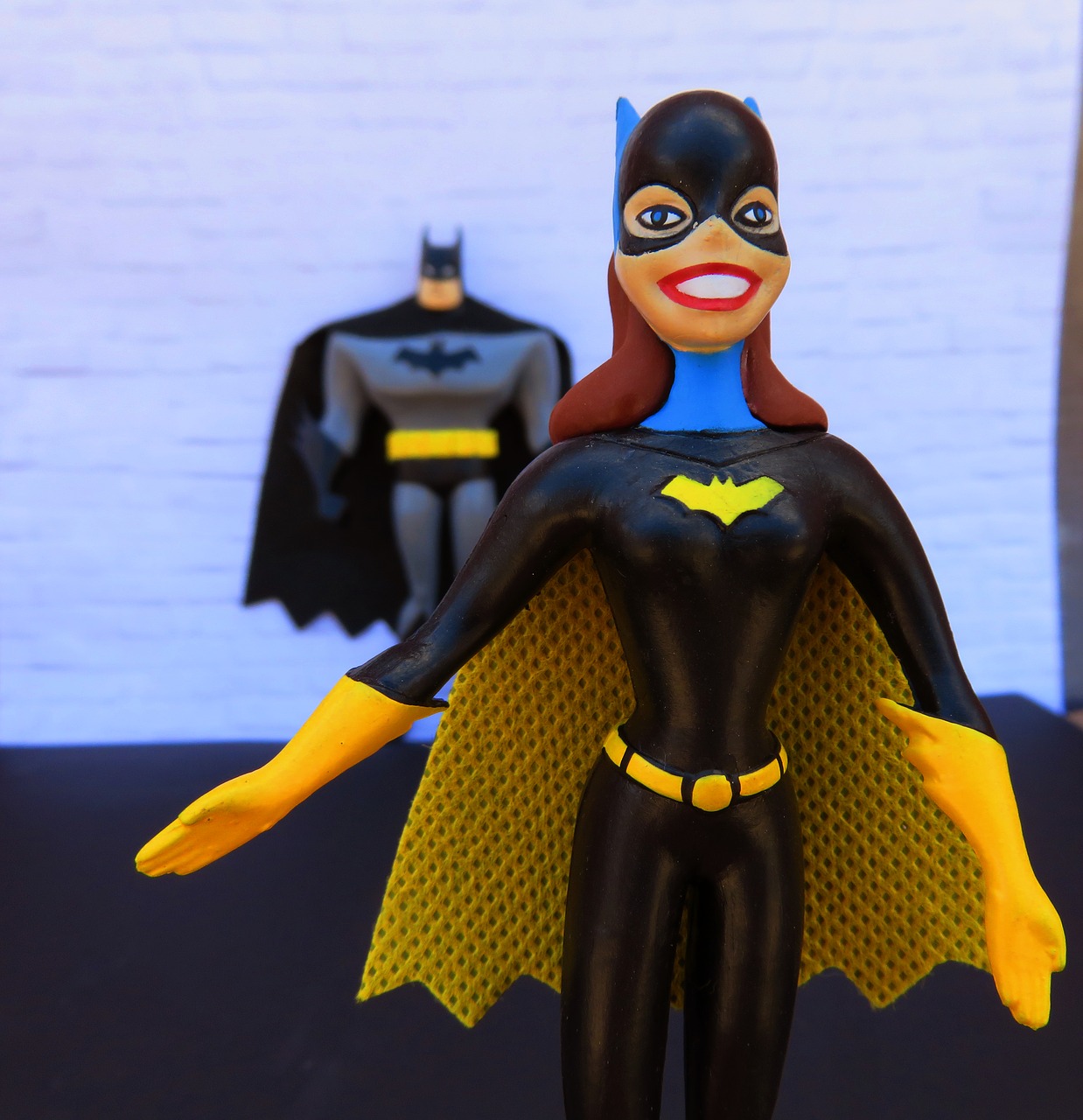 Batgirl-Figur mit breitem Lächeln steht im Vordergrund. Hinter ihr, leicht unscharf, steht Batman an einer weißen Mauer. 