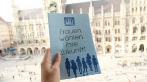 Eine Hand, die einen Plyer hält. Auf dem Flyer steht: Frauen wählen ihre Zukunft. Darüber ist das Logo den Stadtbundes Münchner Frauenverbände gedruckt, darunter die Silhouetten von Frauen. Hinter der Hand mit dem Flyer ist das Münchner Rathaus zu erkennen.