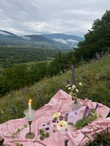 Rosa Decke mit Früchten und Blumen auf einer Bergwiese. Der Ausblick zeigt Wald und Berge. Auf der Decke steht eine brennende Kerze und es ist leicht dämmrig.